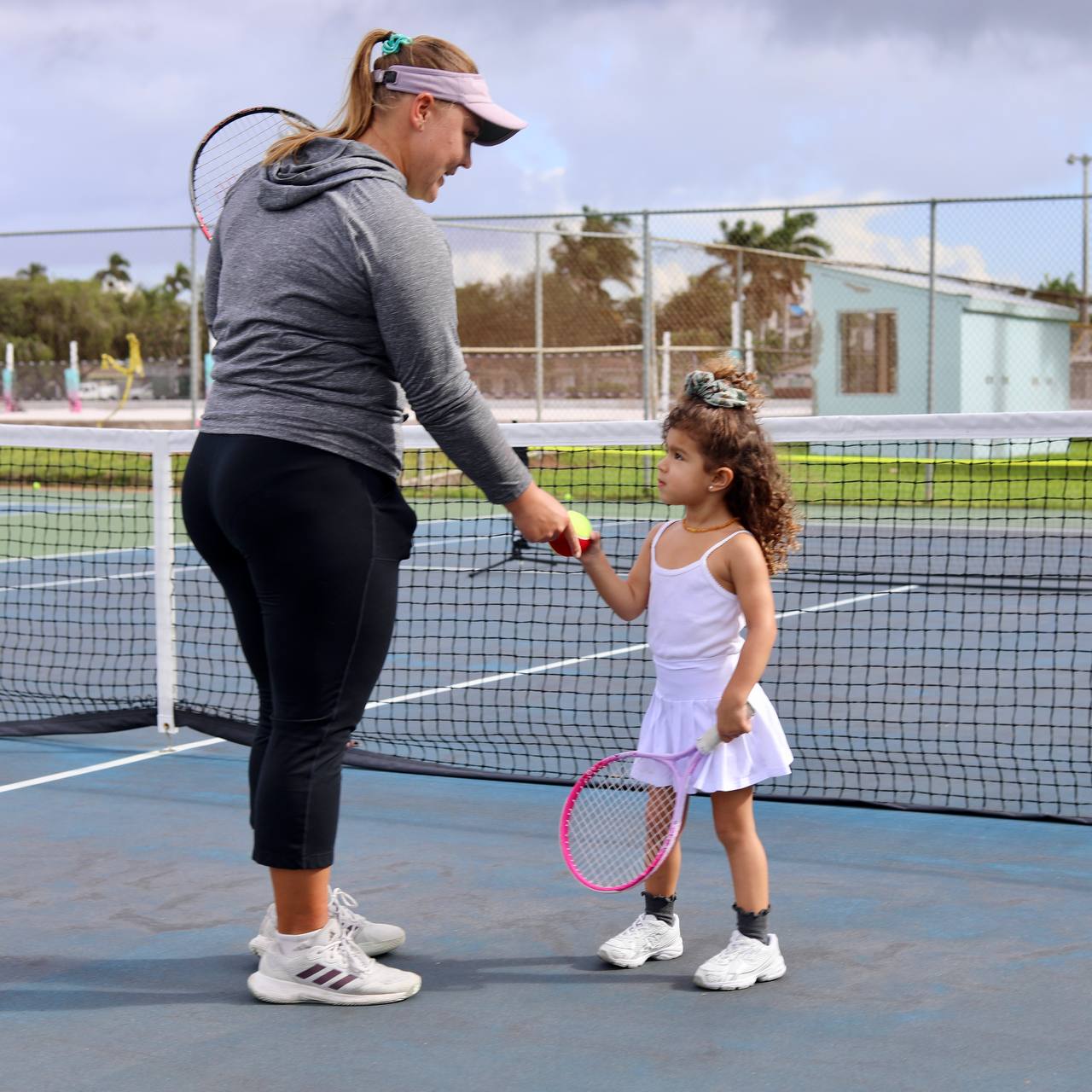 Coach passing a tennis ball to a small girl holding a pink racket